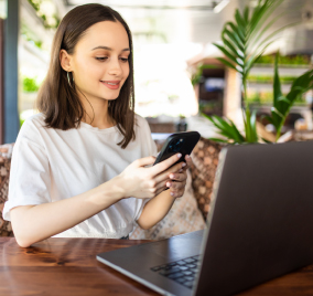 Joven mujer usando su celular frente a un computador portátil en un ambiente moderno y natural.