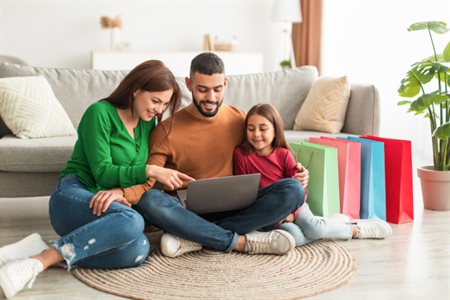 Familia feliz sentada en el piso de su sala de estar, disfrutando de una computadora portátil juntos. El padre, la madre y la hija parecen estar involucrados en una actividad o contenido en la pantalla de la computadora. Todos lucen relajados y cómodos, con expresiones de alegría y concentración. La escena transmite un ambiente acogedor y de conexión familiar, donde comparten una experiencia tecnológica de manera conjunta. Los colores cálidos y el entorno hogareño complementan la sensación de bienestar y disfrute que se aprecia en la imagen.