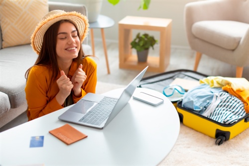 Mujer feliz frente al computador con maleta abierta, lista para viajar gracias a un crédito vacacional.