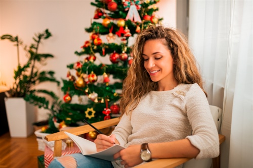 Mujer sonriente escribiendo en un cuaderno frente a un árbol de Navidad, planeando sus compras con un crédito navideño.