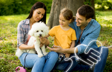 Familia feliz disfrutando de un momento al aire libre con su mascota, una perrita blanca. La madre y el padre están sentados en el césped junto a su hija, sonriendo y acariciando al animal. El entorno es un parque verde y soleado, lo que crea un ambiente acogedor y relajado.