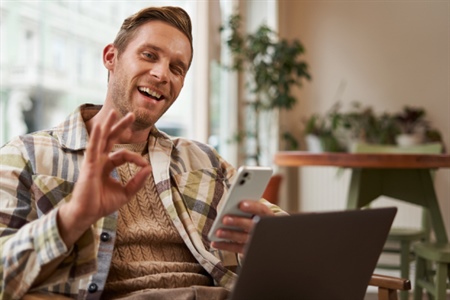 Hombre joven sonriente haciendo gesto de “OK” con la mano, mientras usa su celular y portátil.
