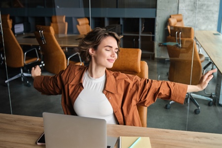 Mujer joven estirándose con expresión de satisfacción en una oficina moderna, frente a su computador portátil.