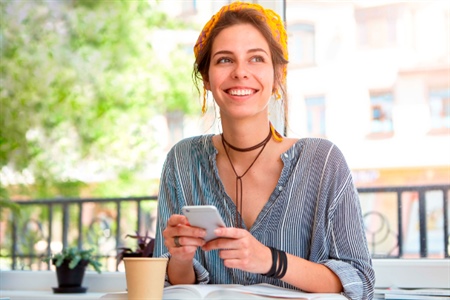Mujer sonriente revisa su celular mientras estudia en una cafetería, ideal para representar la consulta de movimientos financieros desde cualquier lugar.