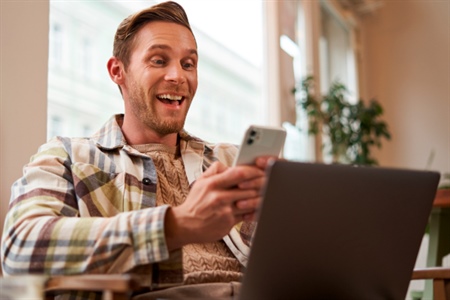 Hombre feliz revisa su celular mientras usa su computador portátil, ideal para ilustrar la consulta de tasas de CDAT de forma digital.